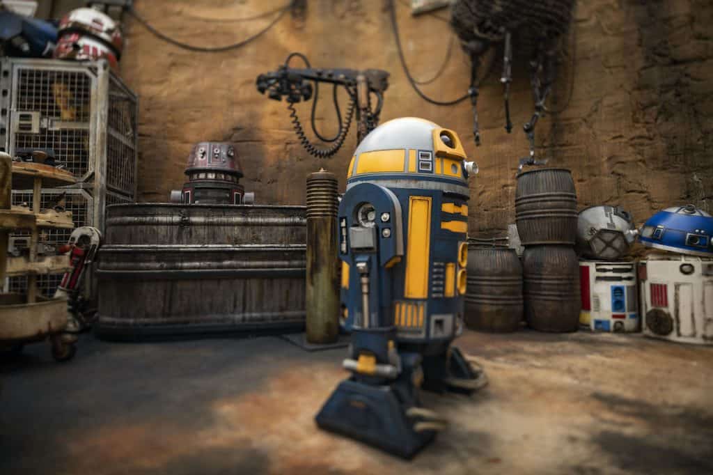A yellow and blue droid at Star Wars: Galaxy’s Edge workshop, surrounded by fellow droids and themed equipment.