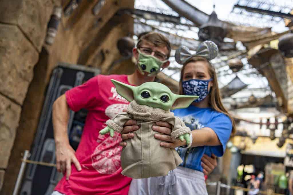 Fans in masks pose with a Baby Yoda plush at Star Wars: Galaxy’s Edge, surrounded by immersive sci-fi theming at Disney’s Hollywood Studios.