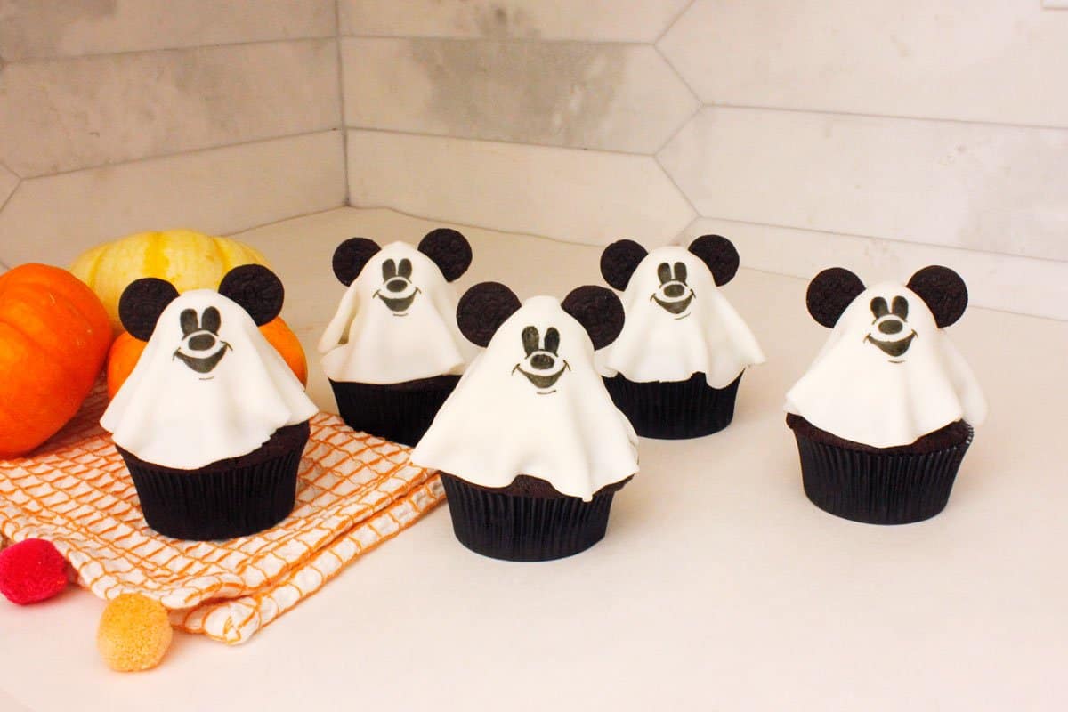 Mickey-themed ghost cupcakes with signature ears await in front of pumpkins and a towel—perfect Halloween magic for Disney fans!.