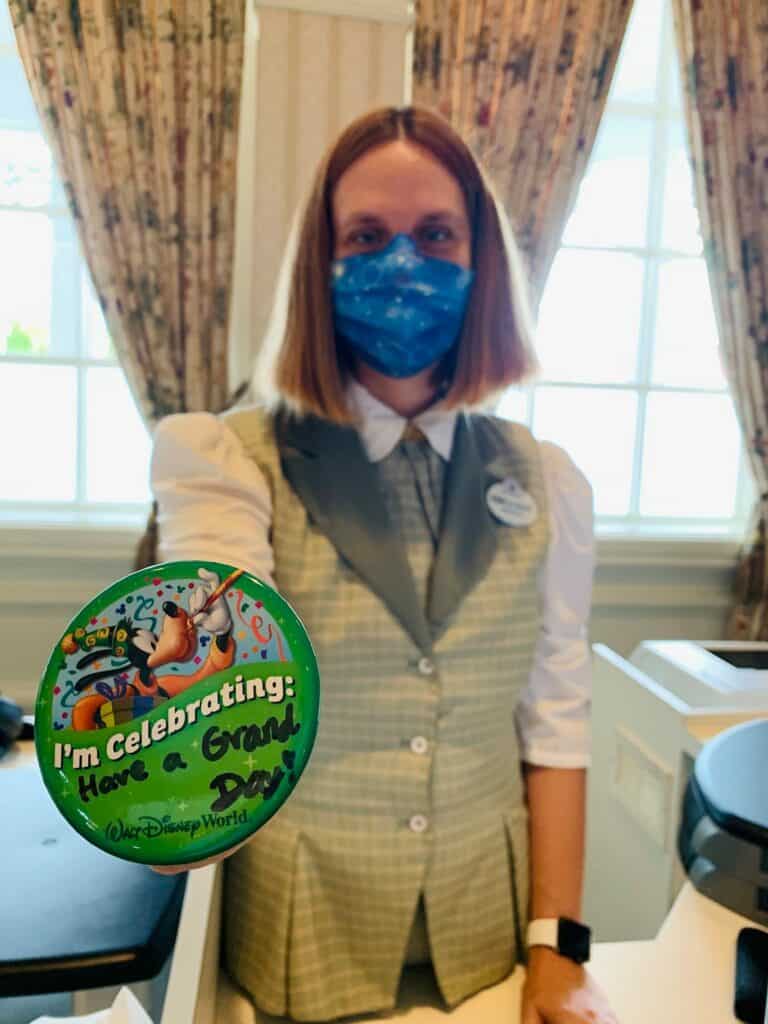 A masked Cast Member at Disney’s Grand Floridian Resort greets guests behind the counter, sporting a celebration button.