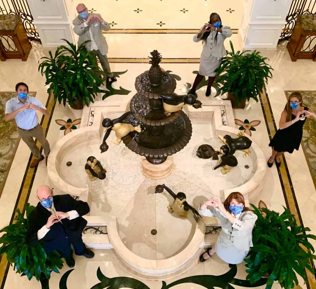 Grand Floridian Cast Members in masks pose by the hotel fountain, making heart signs to share Disney magic with guests.
