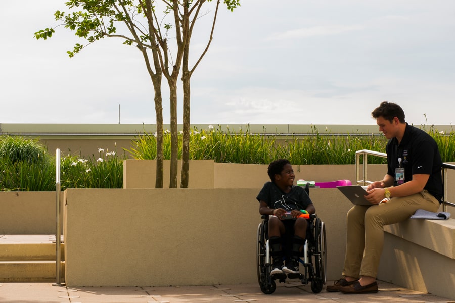 A dad and his son in a wheelchair chat and laugh near a shady tree at the theme park, enjoying togetherness and the outdoors.
