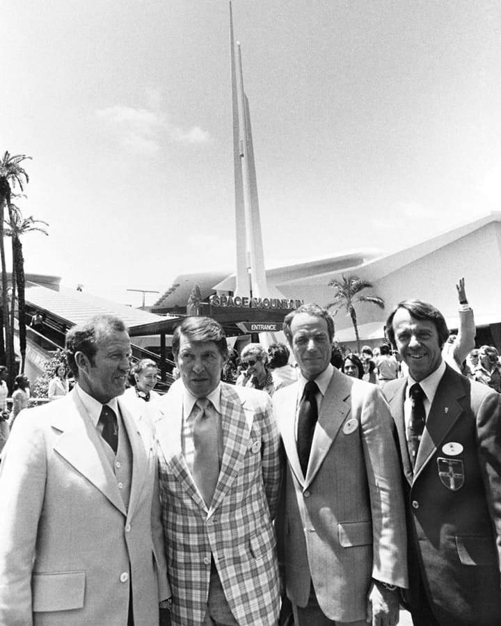 Disney executives in suits celebrate Space Mountain’s opening day at Disneyland, with excited crowds filling Tomorrowland.
