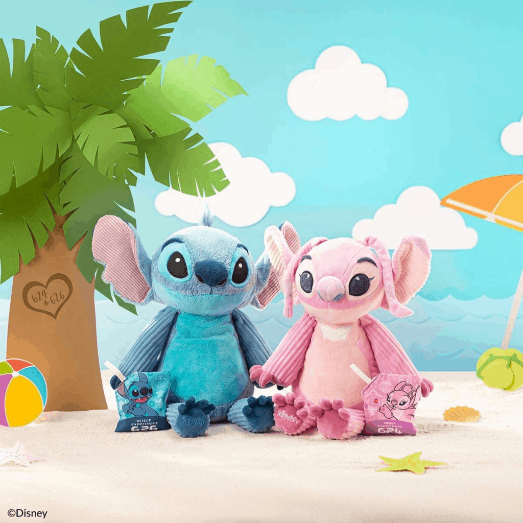 Stitch and Angel plushes from Disney’s Lilo & Stitch relax on a sandy beach scene filled with juice pouches, palm tree décor, and colorful beach toys, bringing the whimsical energy of the Scentsy Collection to life for fans.