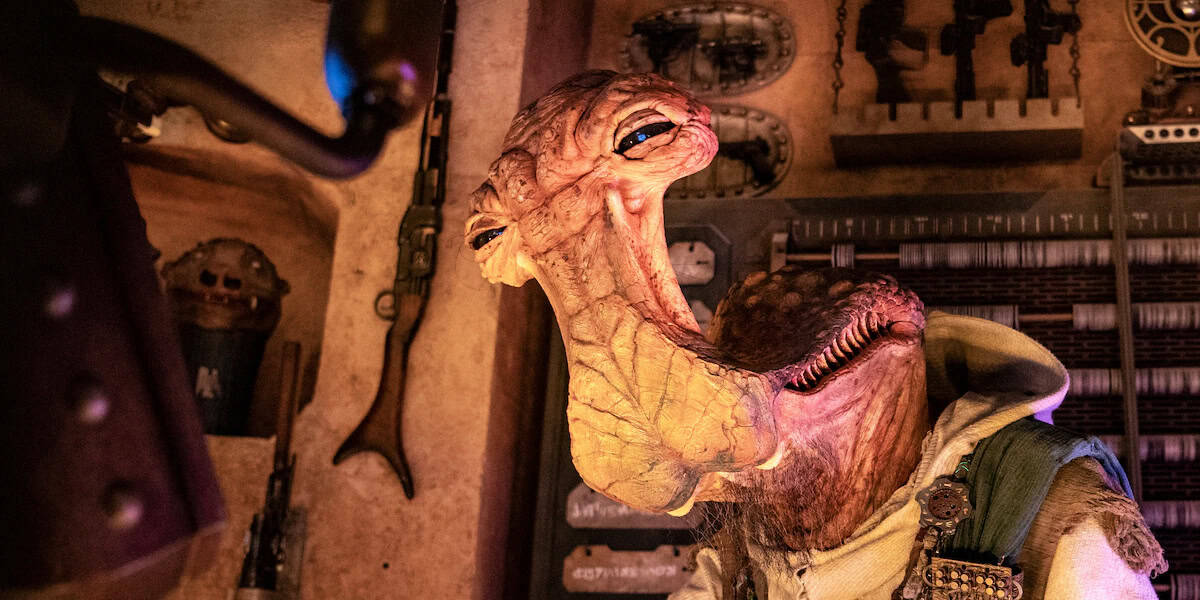 A close-up of an alien animatronic character with wrinkled skin, a long neck, and large blue eyes, standing in a dimly lit, sci-fi themed workshop filled with tools and mechanical parts.