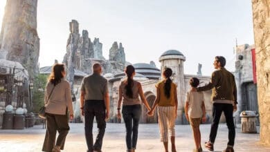 Six people, including adults and children, walk together through a themed area with rocky spires and futuristic buildings, resembling a sci-fi or fantasy setting, likely at an amusement park, viewed from behind.