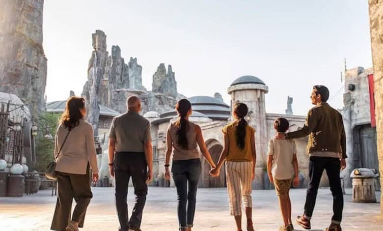Six people, including adults and children, walk together through a themed area with rocky spires and futuristic buildings, resembling a sci-fi or fantasy setting, likely at an amusement park, viewed from behind.