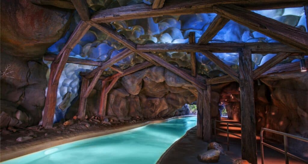 A dimly lit cave-like pool area with rustic wooden beams and a narrow water channel. The ceiling has an undulating, rocky surface, illuminated with blue and purple lighting. A pathway runs alongside the water with a small opening at the end.