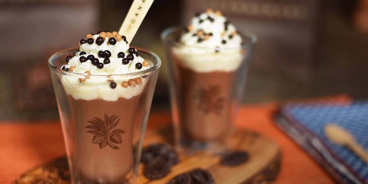 Two glasses of chocolate mousse topped with whipped cream and chocolate pearls evoke the magic of Disney chocolate treats, served on a wooden board with chocolate pieces, a playful stick in one glass, and a blue cloth in the background.