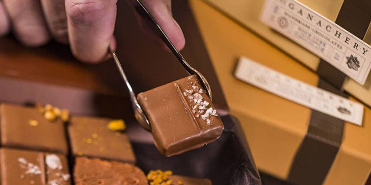 A close-up of a hand using tongs to pick up a square piece of Disney chocolate treat topped with sea salt from a tray of assorted chocolates, with gift boxes in the background.