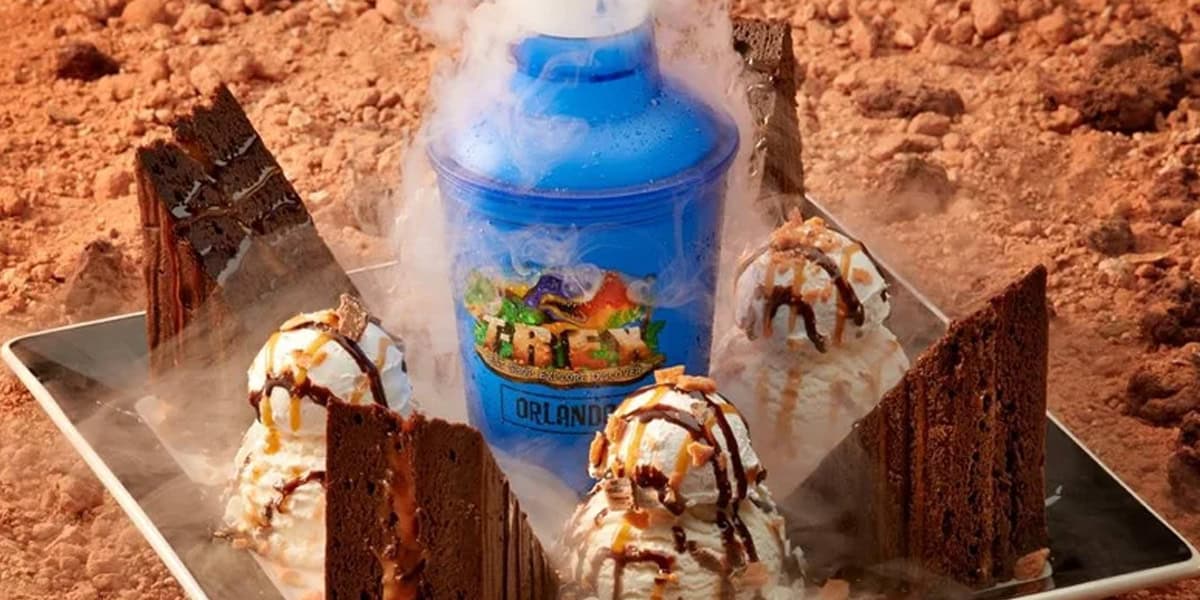 A tray with four scoops of ice cream topped with chocolate and caramel sauce, surrounded by large brownies reminiscent of Disney chocolate treats, with a blue T-Rex Orlando souvenir cup in the center, all set on smoky dry ice.