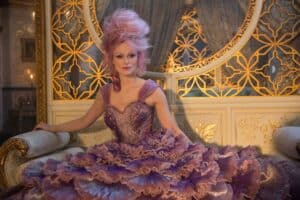 Keira Knightley with elaborate pink hair and a lavender, voluminous gown adorned with ruffles sits gracefully on a luxurious, ornate couch. The background features intricate golden designs and a large, circular mirror reflecting an opulent room.