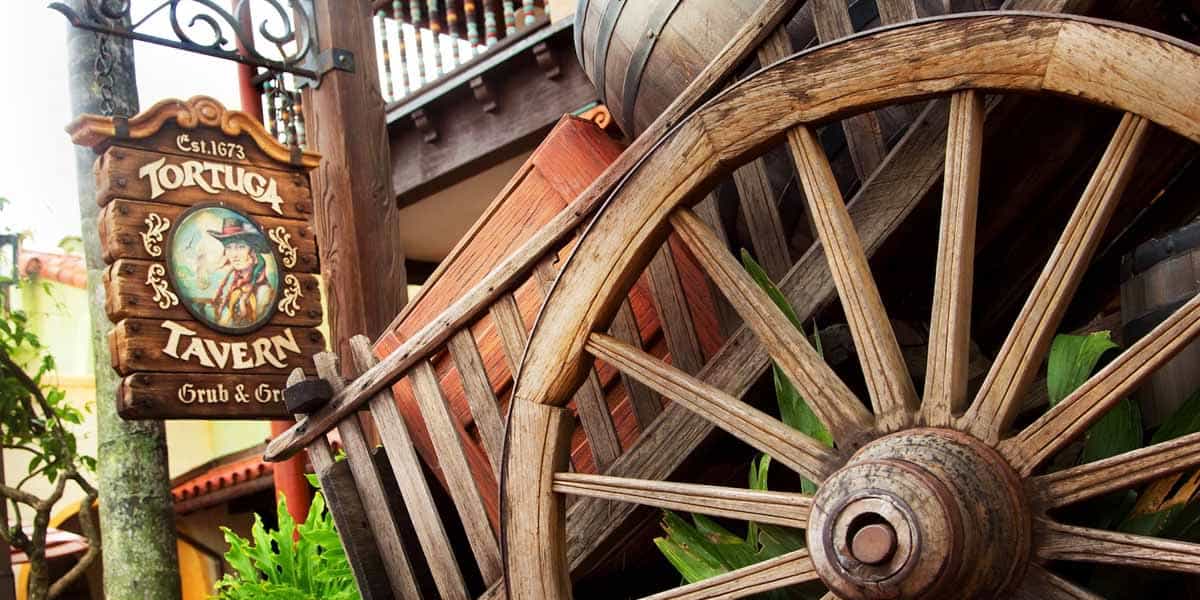 A rustic wooden sign reading “Tortuga Tavern Grub & Grog” hangs next to a large wagon wheel and barrels, inviting guests to enjoy Adventureland dining amid tropical plants and a charming building in the background.