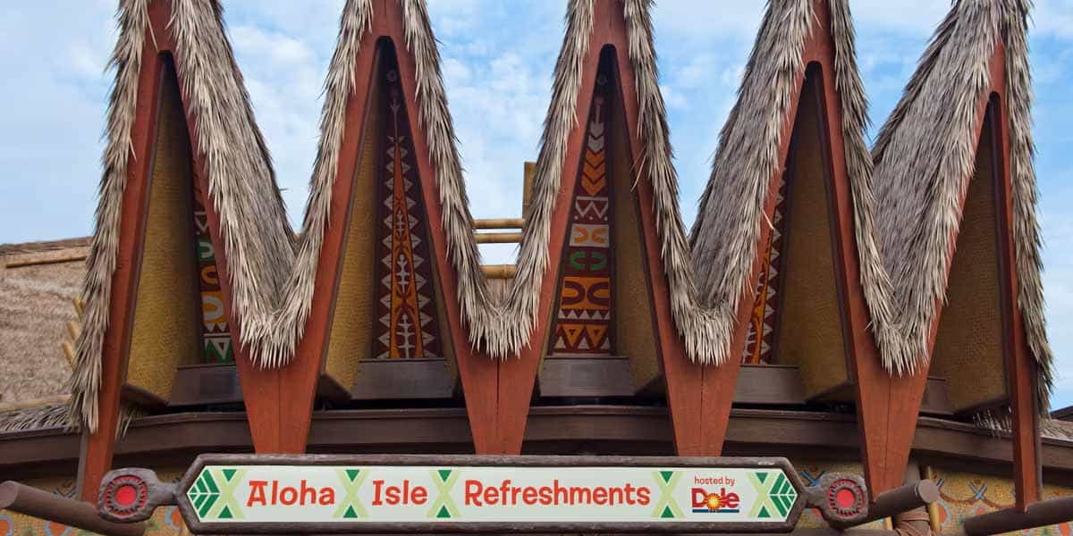 A building with a thatched roof and decorative, pointed wooden peaks displays a sign reading "Aloha Isle Refreshments" with colorful patterns—a popular Adventureland dining spot hosted by Dole.