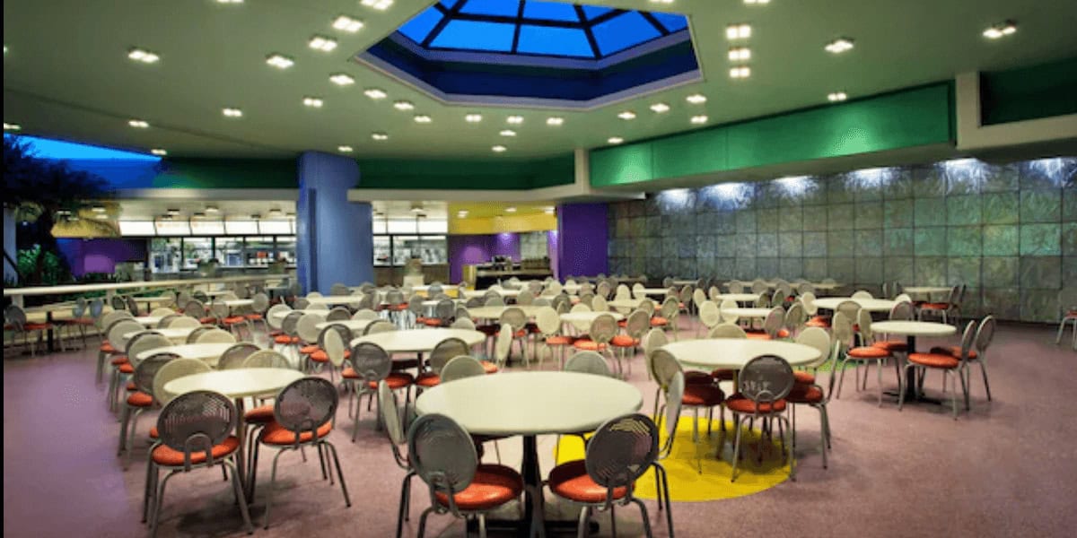 A brightly lit, colorful cafeteria with round tables and red chairs sits under a large skylight—one of those secret spots in Magic Kingdom. The empty space features a food counter at the back and vibrant green, purple, and yellow walls.