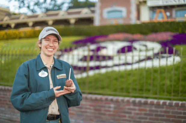 A cheerful Disneyland Cast Member in uniform presents a chocolate-covered apple, standing in front of the vibrant Minnie Mouse floral display on Main Street, U.S.A.