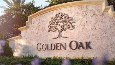 A stone sign with an ornate tree design and the words "GOLDEN OAK" engraved on it, surrounded by greenery and purple flowers, sunlight shining from the left—perfect for those moving close to Disney’s magical bubble.