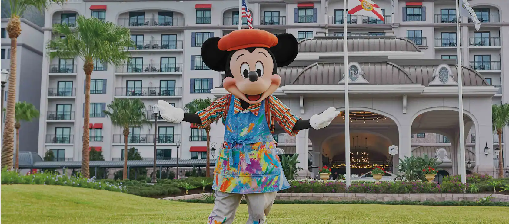Mickey Mouse, in a colorful painter outfit, stands cheerfully on a lawn in front of a large resort building with balconies. Nearby, palm trees sway by three flagpoles, all set to welcome the exciting new DVC lounge at Disney World.