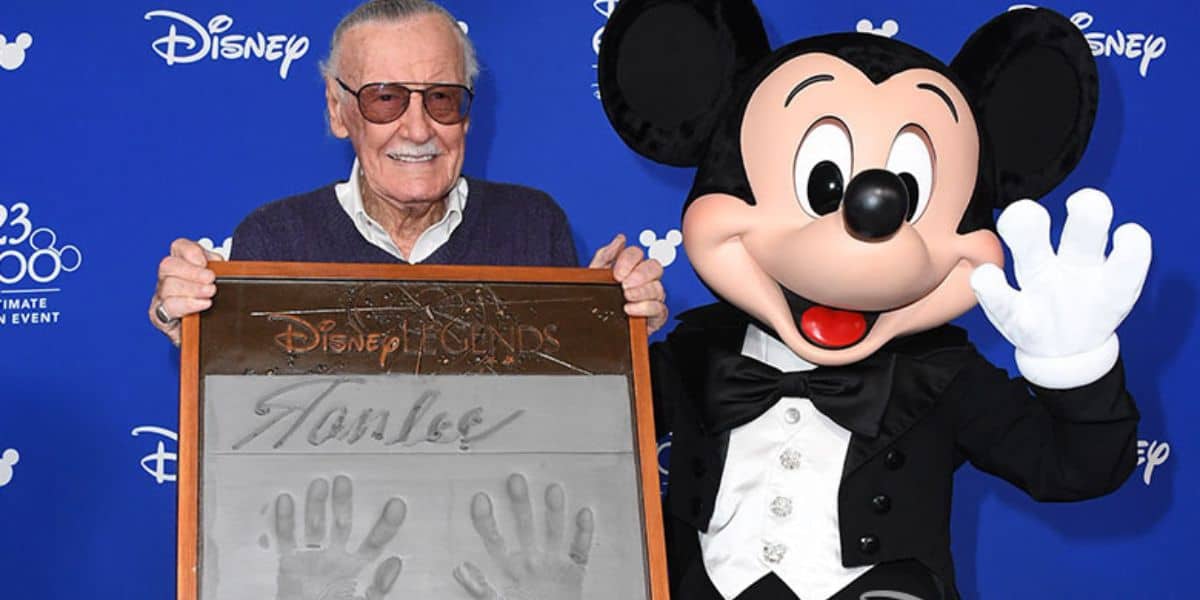 Stan Lee smiles while holding a framed plaque of his handprints, standing next to Mickey Mouse in a tuxedo, against a blue Disney Legends event backdrop—an iconic moment for any Stan Lee documentary or Jack Kirby fan.