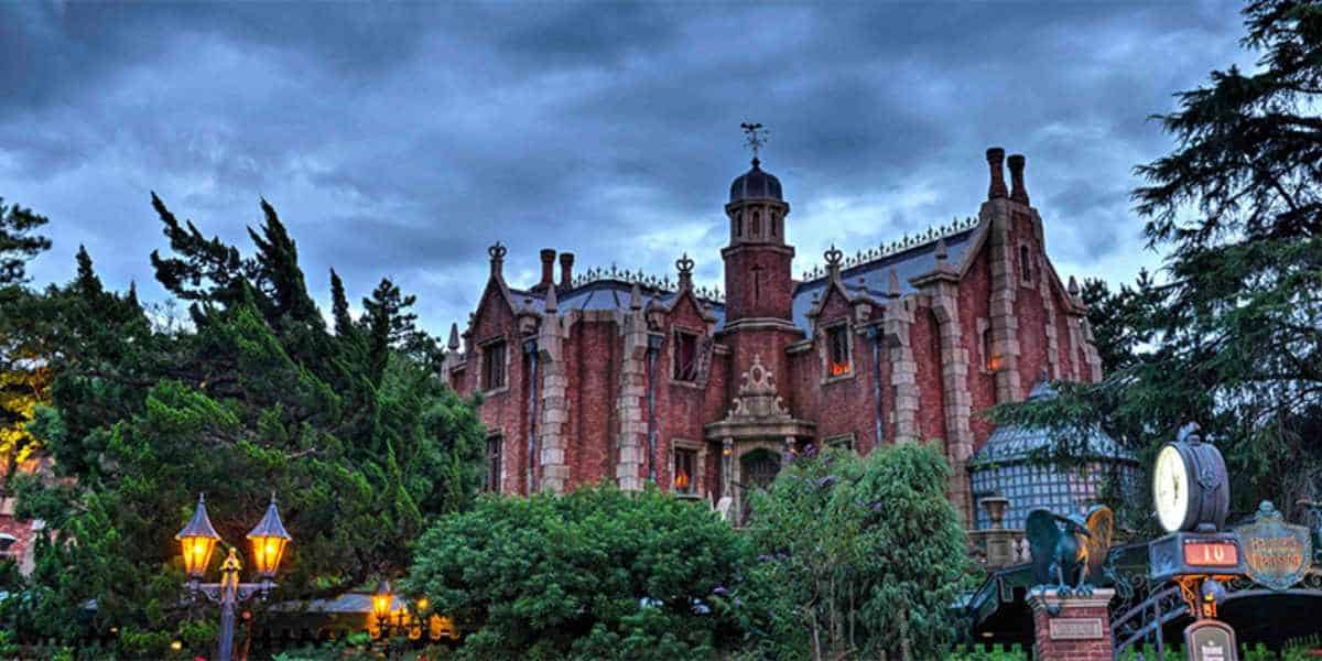 A large, gothic-style brick mansion with towers and ornate details stands surrounded by lush greenery under a cloudy, twilight sky. Vintage lamps and signs are visible in the foreground, hinting at exclusive entry—like holding a Tokyo Disney Priority Pass.