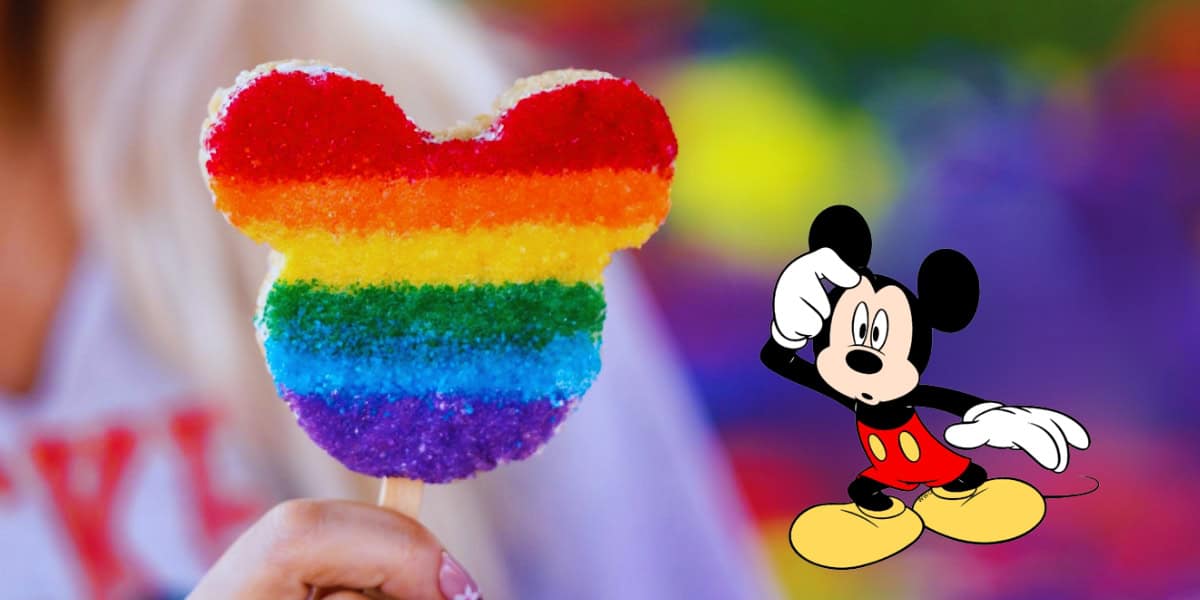 A person holds a rainbow-colored sugar cookie shaped like Mickey Mouse on a stick. The background is blurred with colorful lights, and an image of Mickey Mouse in his classic red shorts and yellow shoes is positioned on the right side of the image, subtly hinting at Disney's ongoing attempts at LGBTQ representation.