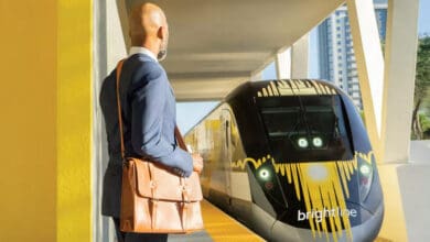 A man in a suit with a brown shoulder bag waits on a yellow train platform as a modern Brightline train, known for its Disney World transportation, approaches under the sunny, covered station heading to Disney World.
