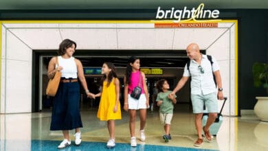 A smiling family of five walks hand in hand through a Brightline train station, with two parents and three children carrying bags, ready for their Disney World adventure. The station sign above them reads "Brightline" and "Orlando Health near Disney World.