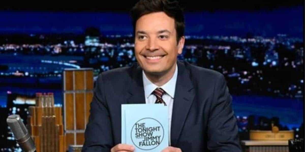 Jimmy Fallon in a suit smiles while holding a card emblazoned with "Jimmy Fallon" and "The Tonight Show Starring Jimmy Fallon." He is seated at a desk with a microphone, the vibrant cityscape serving as his backdrop.