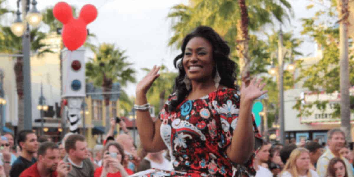 A woman with a joyful expression, wearing a patterned red dress, waves from a parade float; a large crowd and mickey mouse-shaped balloons are in the background.