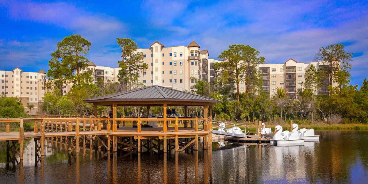 A lakeside pavilion with a wooden dock extends over the water. Nearby, pedal boats shaped like swans are moored. In the background, several mid-rise buildings are partially obscured by trees. The sky is clear and blue, indicating fair weather.
