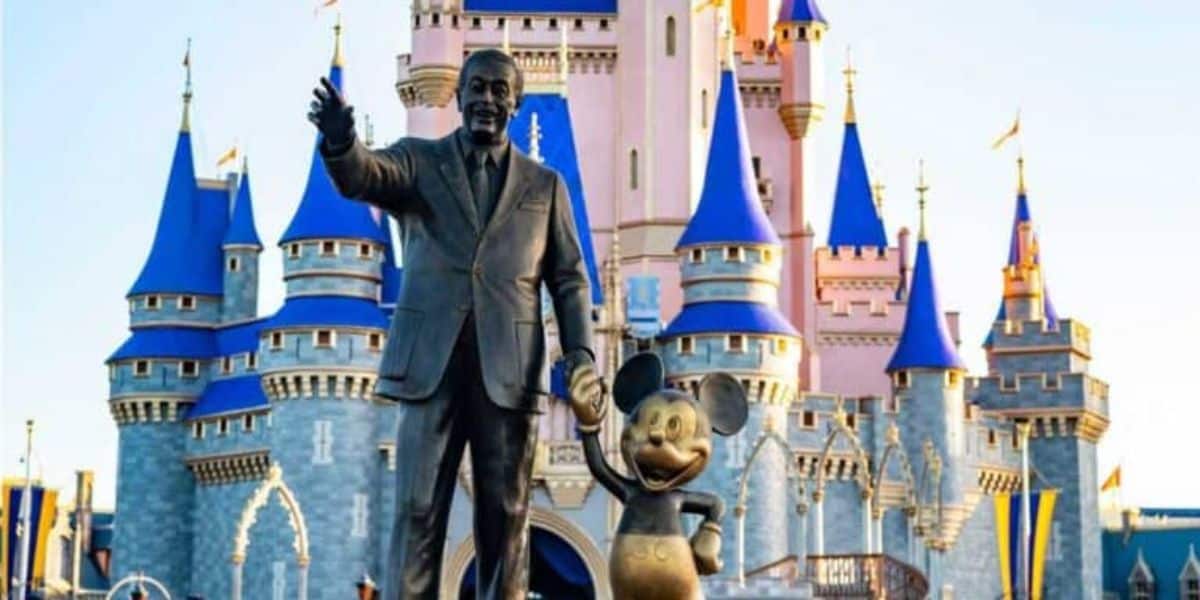 A statue of a man holding hands with a cartoon mouse, symbolizing Disney's legacy amidst ongoing discussions about corporate greed, stands in front of a grand castle with blue conical roofs and pink walls, under a clear blue sky.