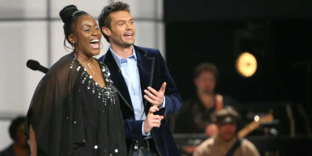 A woman and a man stand together on a stage, smiling and speaking. The woman is wearing a black, embellished outfit, and the man is in a dark blazer over a light shirt. A microphone is visible to the left, with musicians blurred in the background.