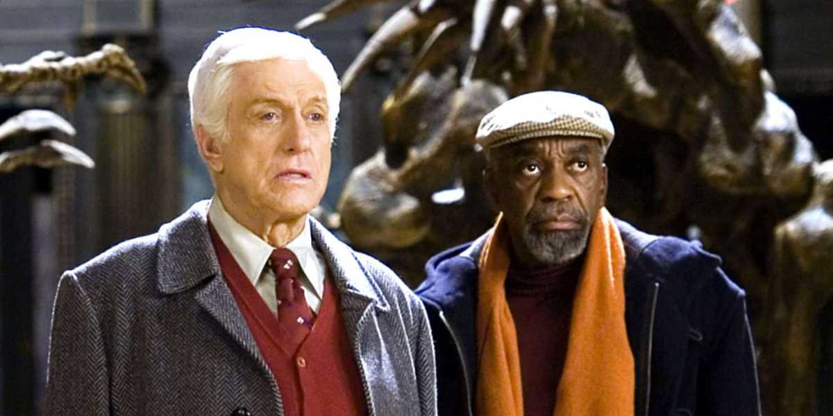 Two older men stand side by side indoors. The man on the left wears a gray coat, a red tie, and a white shirt, while the man on the right wears a black jacket, an orange scarf, and a beige cap. They appear to be in a museum or exhibit with large dinosaur statues behind them.
