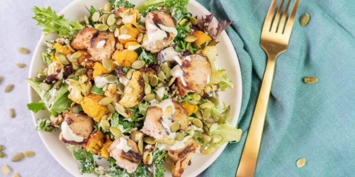 A white plate filled with a mixed greens salad topped with chunks of grilled chicken, roasted cauliflower, seeds, and a creamy dressing. A gold fork is placed to the side of the plate, with a green napkin underneath them on a light-colored surface.