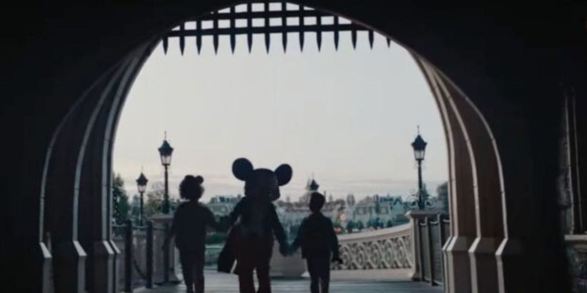 Silhouettes of three characters, including one with large mouse ears, walk hand in hand under a stone archway at Disneyland Paris Resort. Iron gates are above, while lamp posts line the curved bridge leading to a distant landscape.