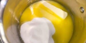 A metal saucepan containing a partially melted stick of butter and a mound of white granulated sugar.