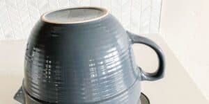 A dark gray ceramic bowl with a handle is placed upside down on a light-colored countertop. In the background, there is a white, textured backsplash with a geometric pattern. The bowl has a glossy finish and visible ridges.