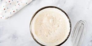 A glass bowl filled with frothy, mixed batter is placed on a white marble countertop. Beside the bowl, there is a metal whisk. A section of a white kitchen towel with red and blue star patterns is partially visible in the top left corner.