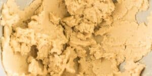 Close-up of creamy, beige cookie dough in a mixing bowl. The dough is unevenly spread with a slightly coarse texture, hinting at its thick and rich consistency.