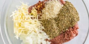A glass bowl filled with ground meat and various seasonings including shredded cheese, chopped onions, breadcrumbs, red chili flakes, and a large portion of dried herbs. The ingredients are arranged in separate piles on top of the meat.