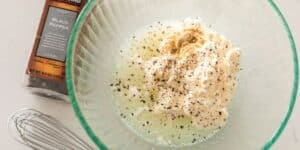 A transparent bowl contains a mixture of mayonnaise, spices, and lemon juice being prepared. Next to the bowl is a container of black pepper and a metal whisk, indicating the ingredients being used for the recipe.