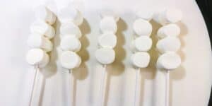 Six sticks with clusters of marshmallows on a white surface. Each stick has four marshmallows, arranged vertically in a row.