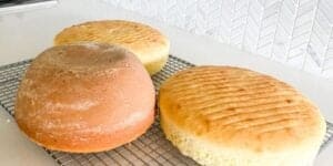 Three round loaves of bread cooling on a wire rack on a white countertop. The bread in the foreground is darker and crustier, while the other two loaves are lighter in color with a smooth, slightly impressed pattern on top. The background features white herringbone tiles.