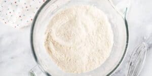 A glass mixing bowl filled with flour sits on a marble countertop next to a metal whisk. A white cloth with red and blue stars is partially visible in the background.
