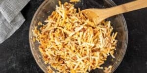 A clear glass bowl holds a mixture of shredded cheese and bacon bits. A wooden spoon rests in the bowl, ready for stirring. The bowl is placed on a dark textured surface, with a white and black checkered cloth visible in the top left corner of the image.