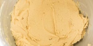 A close-up image of a stainless steel mixing bowl filled with smooth, creamy cookie dough. The dough looks well-mixed and ready for baking, with a pale yellow color and a slightly sticky texture.