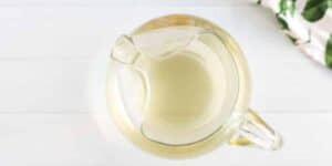 A top-down view of a glass pitcher filled with a light, transparent liquid, likely lemonade or another beverage, placed on a white surface. Part of a green and white cloth is seen in the upper right corner.