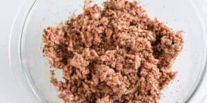A clear glass bowl containing a mixture of ground beef, finely chopped onions, and herbs. The meat is seasoned and appears ready for cooking, set against a light background.