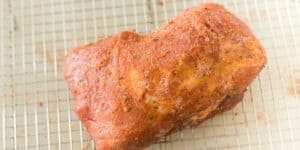 A piece of seasoned raw pork roast sits on a wire rack over a baking sheet. The meat is coated with a mix of spices, including visible flecks of herbs and coarse salt.