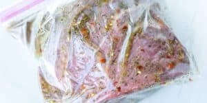 A piece of raw meat is marinating in a clear, ziplock plastic bag. The marinade contains various herbs, spices, and red pepper flakes, creating a speckled appearance on the meat. The bag is sealed and resting on a white surface.